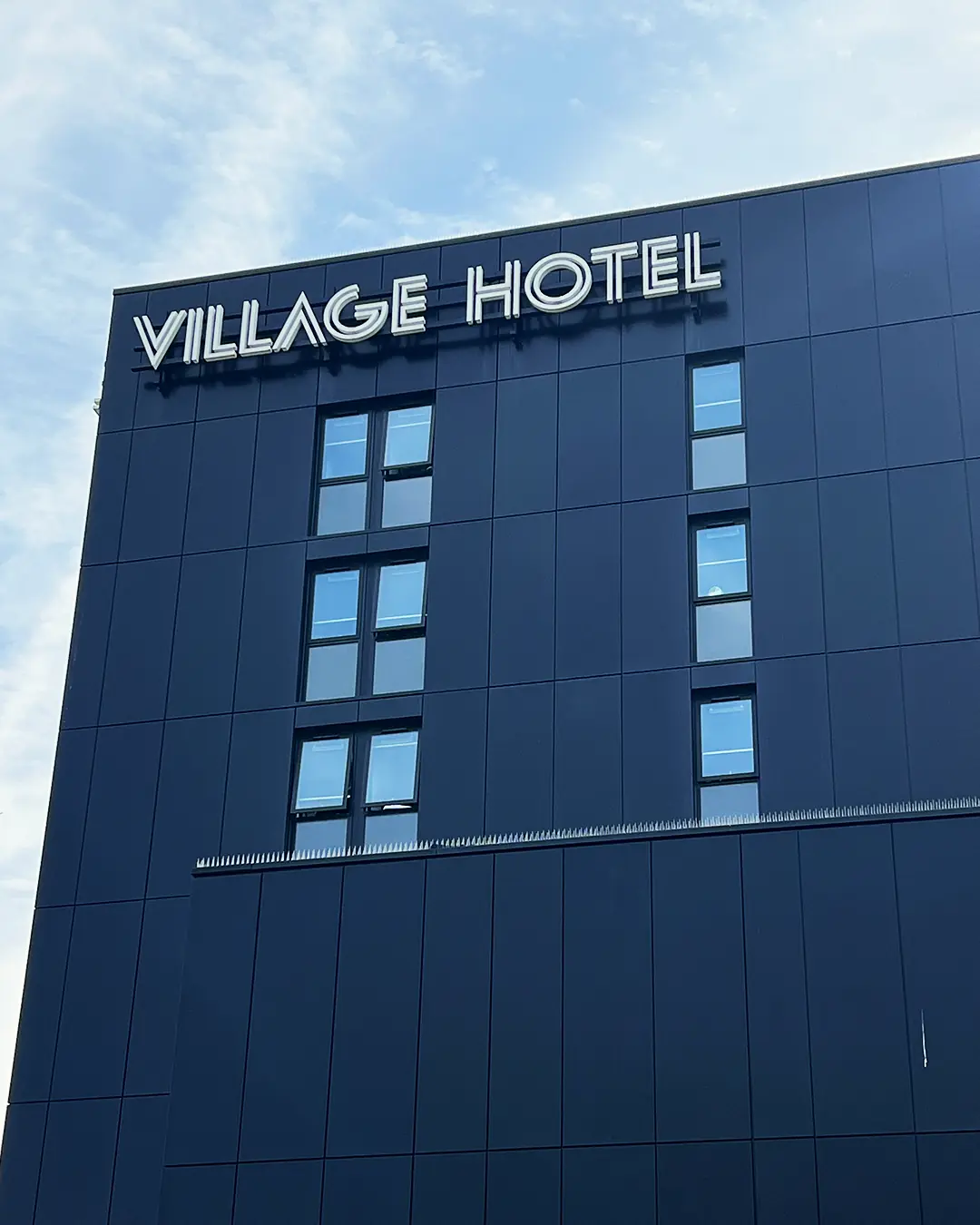 Built-up letters reading VILLAGE HOTEL on the dark clad facade of a hotel