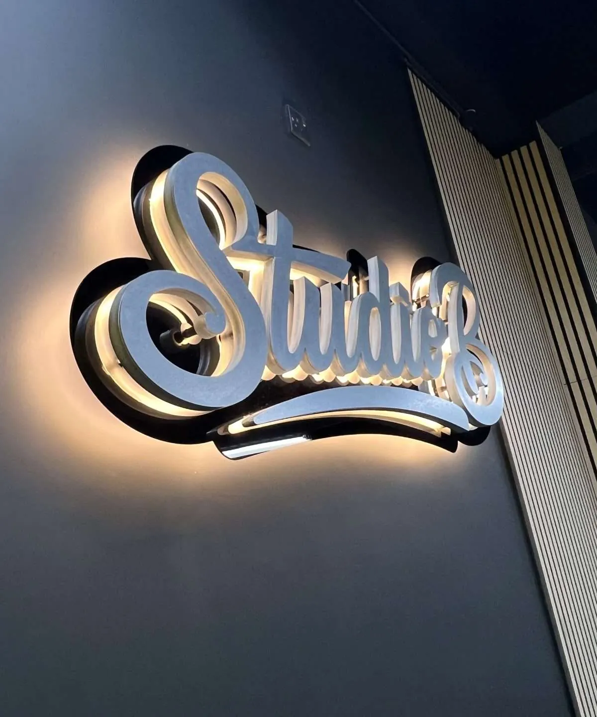 Studio B sign with its halo on, on a dark wall between timber slats