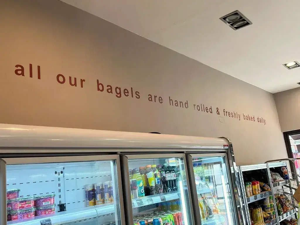 Lettering along the wall above the fridges inside Fress Bagelry: all our bagels are hand rolled and freshly baked daily