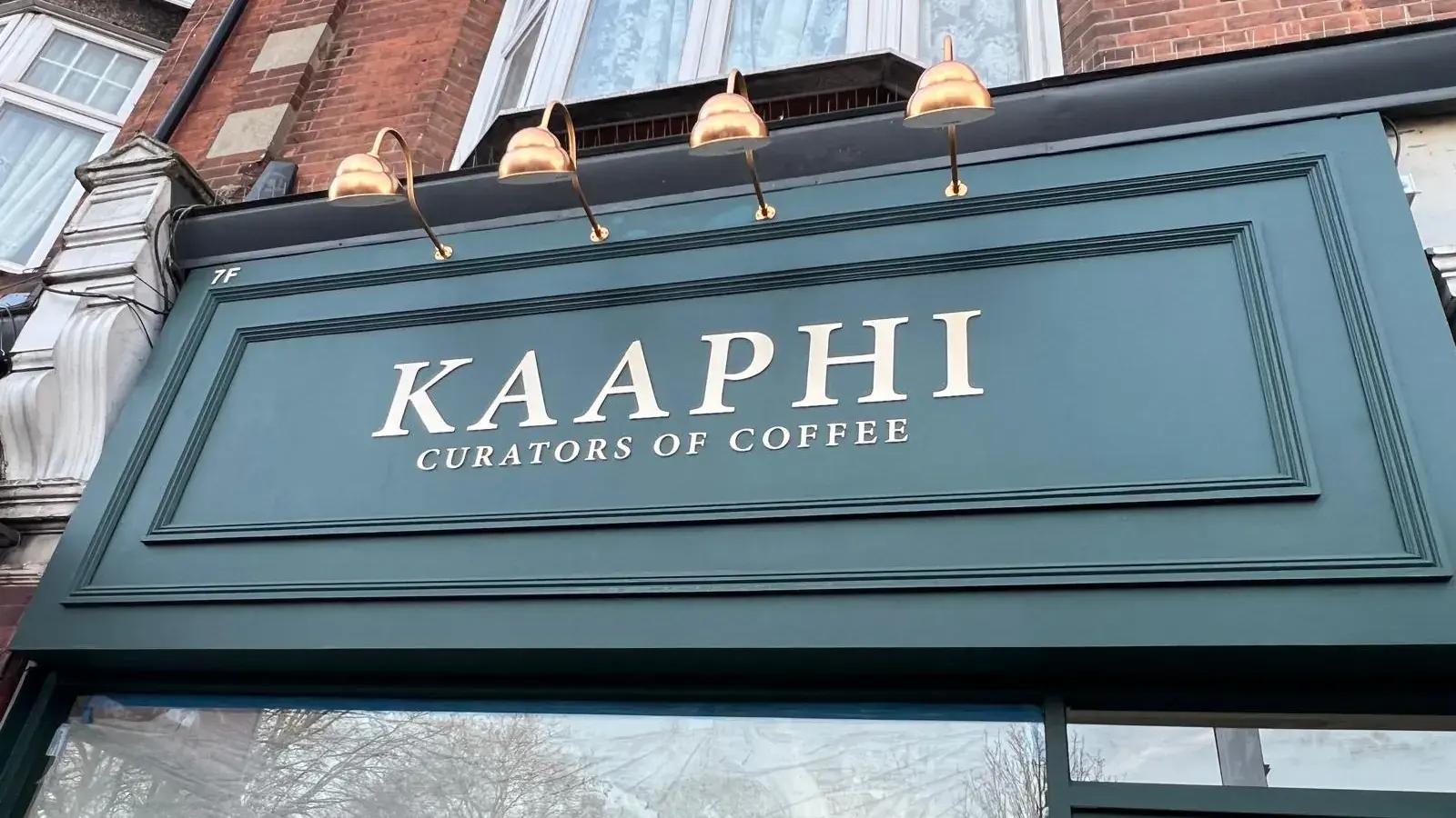 The Kaaphi fascia from below: raised gold-finish letters reading Kaaphi, Curators of Coffee, inside the painted moulded panel frame, with swan-neck lights above