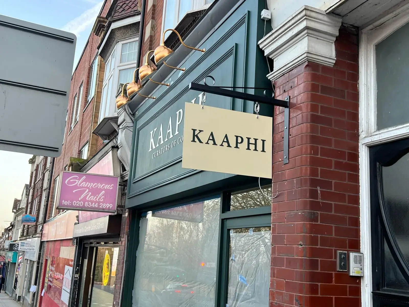 Cream Kaaphi hanging sign on a black bracket fixed to the brick pier beside the shop front