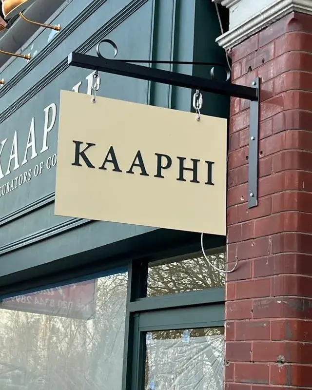 Cream hanging sign lettered Kaaphi in black, hung from a black bracket on the brick pier beside the fascia