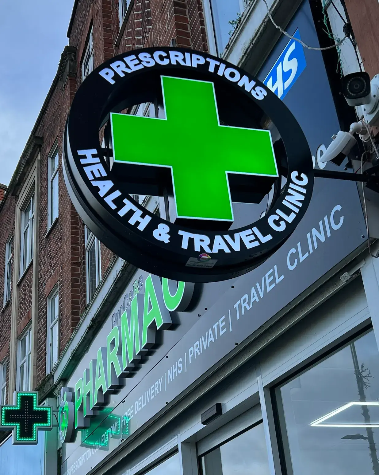 Round illuminated projecting sign with a green pharmacy cross at ProCare Pharmacy, lettered Prescriptions and Health & Travel Clinic