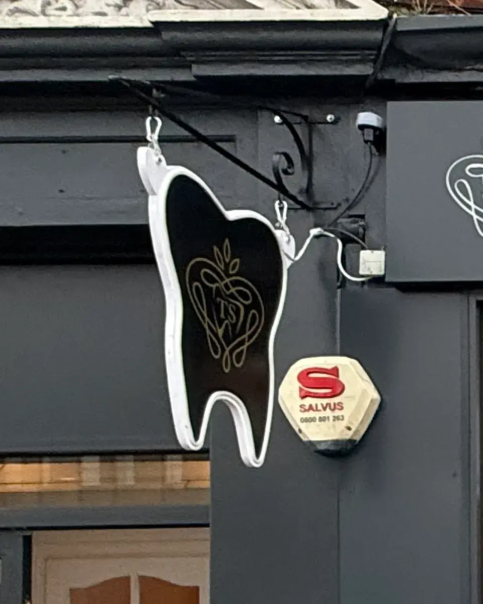 Tooth-shaped hanging projecting sign in black and gold on a scroll bracket at Tigga Smile Bedford