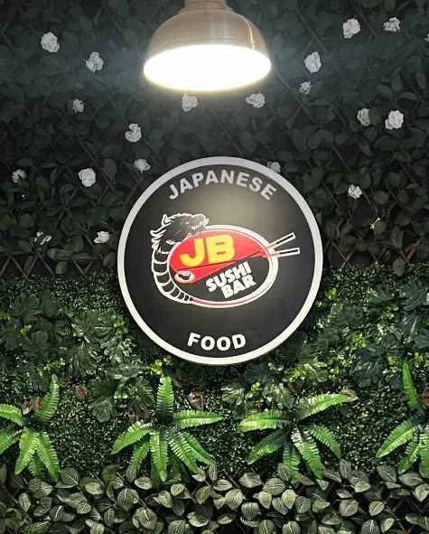 Round JB Sushi Bar sign on a wall of artificial leaves under a pendant lamp